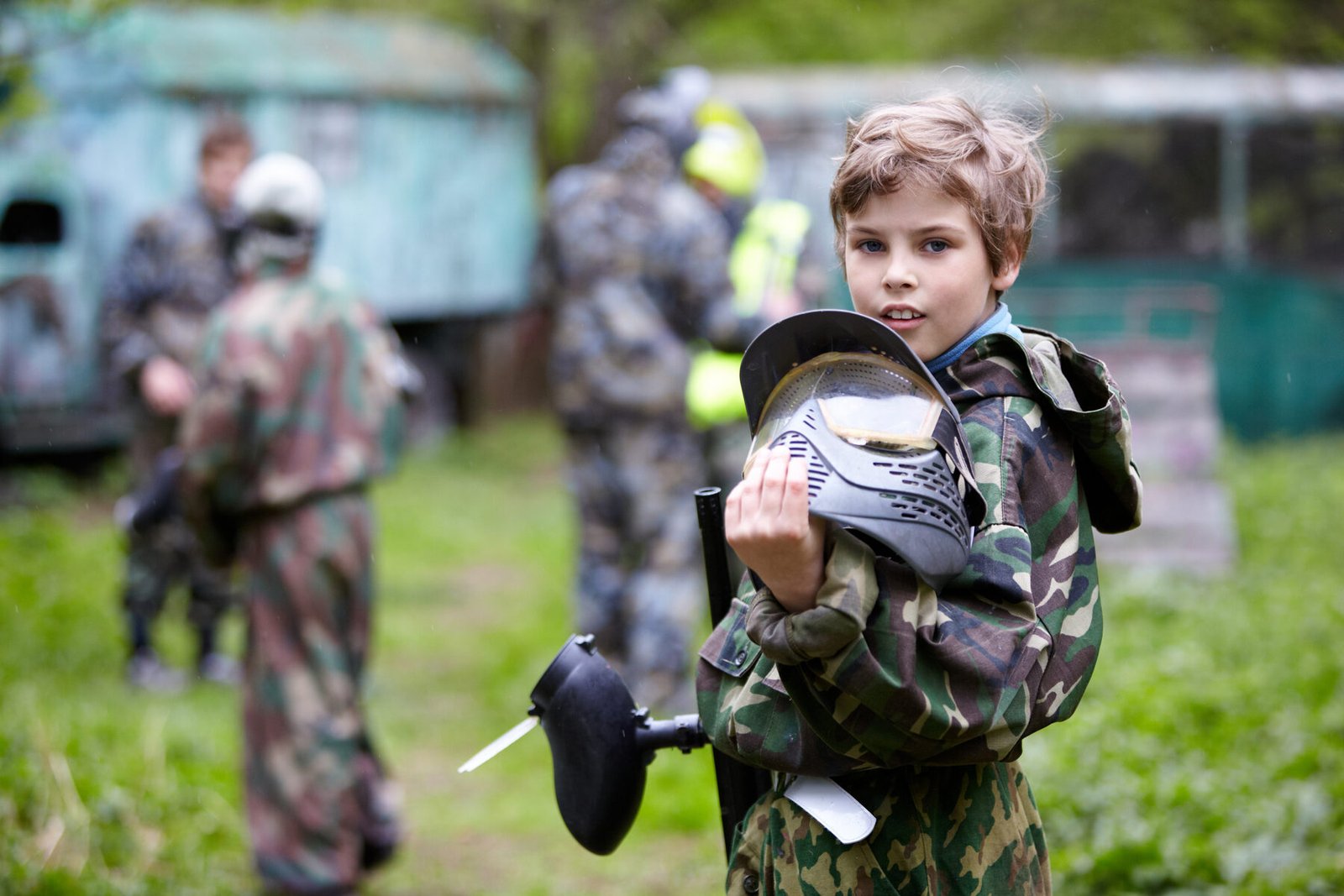 A young player in camouflage holding a paintball mask and marker at an outdoor field